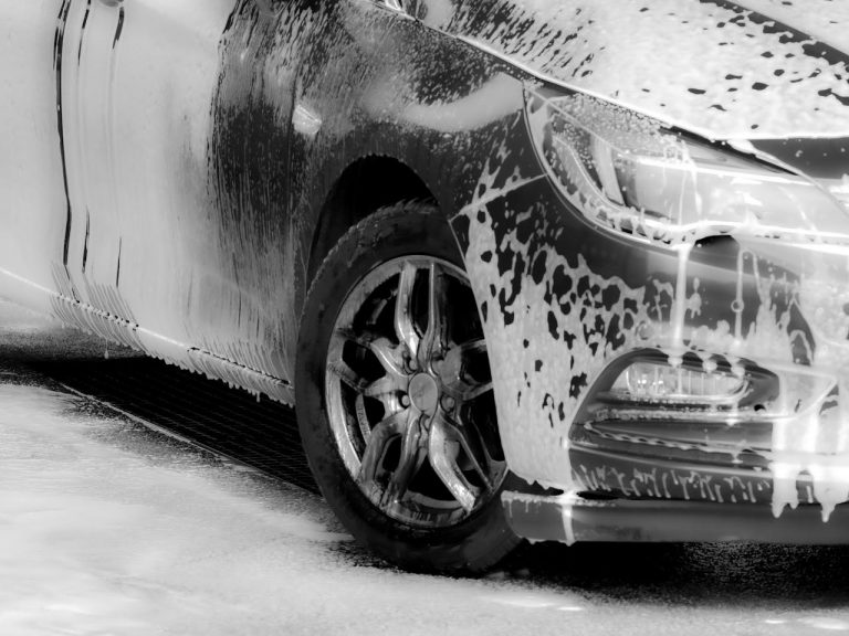 Black and white photo of a car wheel and body covered in soap foam during a wash.